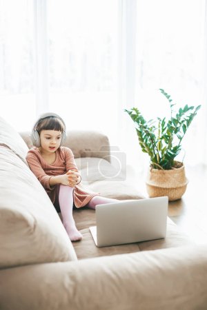 Téléchargez les photos : Une douce fille de 5-6 ans portant un casque tout en regardant un ordinateur portable, assis sur un canapé. Concept : relaxation induite par la technologie, éducation en ligne, divertissement technologique - en image libre de droit