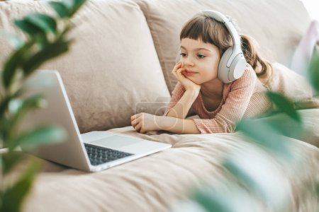 Téléchargez les photos : Une charmante fille de 5-6 ans allongée sur un canapé, portant un casque et regardant un écran d'ordinateur portable. Concept : relaxation imprégnée de technologie, éducation en ligne, divertissement informatique - en image libre de droit