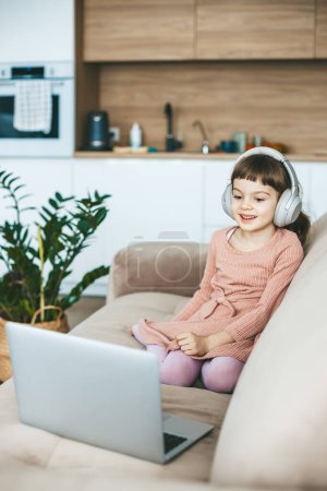 Téléchargez les photos : Une jolie petite fille souriante regardant un écran d'ordinateur portable, assise sur un confortable canapé beige. Concept : relaxation imprégnée de technologie, éducation en ligne, divertissement informatique - en image libre de droit