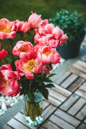 Un beau bouquet de pivoines roses. Ils sont debout dans un vase en verre clair sur une terrasse en bois