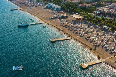 Téléchargez les photos : Vue aérienne du front de mer d'une station balnéaire avec des eaux turquoises claires et des jetées en bois. De nombreux parasols et chaises longues blancs sont éparpillés sur la plage de sable fin - en image libre de droit