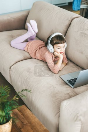 Téléchargez les photos : Détente sur un canapé moelleux, une gentille petite fille portant des écouteurs alors qu'elle est absorbée par son écran d'ordinateur portable. Concept : relaxation imprégnée de technologie, éducation en ligne, divertissement informatique - en image libre de droit