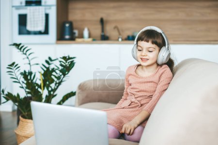 Téléchargez les photos : Une fillette souriante de 5-6 ans regardant un écran d'ordinateur portable, assise sur un canapé beige. Concept : relaxation imprégnée de technologie, éducation en ligne, divertissement informatique - en image libre de droit