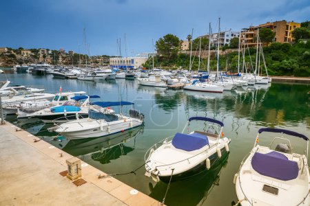 Téléchargez les photos : Le port de Porto Cristo à Majorque offre une scène sereine avec des bateaux doucement ancrés dans un contexte de bâtiments côtiers et de verdure luxuriante - en image libre de droit