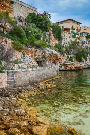 Das Bild der Küste in Porto Cristo, Mallorca, zeigt ruhiges türkisfarbenes Wasser vor felsigem Hintergrund, mit Gebäuden, die in die Landschaft eingebettet sind