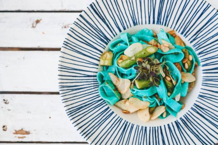 Téléchargez les photos : Une assiette de pâtes bleues végétariennes vibrantes, garnie d'asperges fraîches, de pignons de pin et de légumes verts, est servie dans une assiette blanche ornée de rayures bleues. Vue du dessus, espace de copie - en image libre de droit