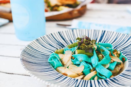 Téléchargez les photos : Des pâtes bleu végétarien éclatantes, rehaussées d'asperges, de pignons de pin et de verts, sont élégamment présentées dans une assiette blanche ornée de rayures bleues. - en image libre de droit