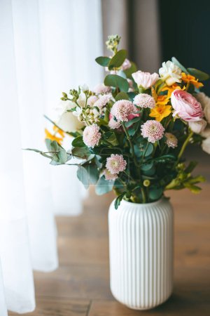 Ein lebhafter bunter Strauß aus verschiedenen Blumen in einer weißen, gerippten Vase, die auf einem Holzboden steht, mit dem weichen natürlichen Licht aus dem Fenster