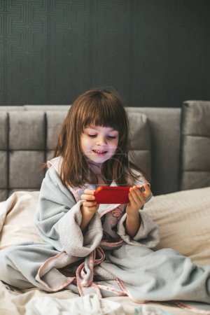 Téléchargez les photos : Une jeune fille portant un peignoir assis sur un lit, absorbée par le contenu numérique avec son smartphone dans les mains. Concept : enfants et smartphones - en image libre de droit