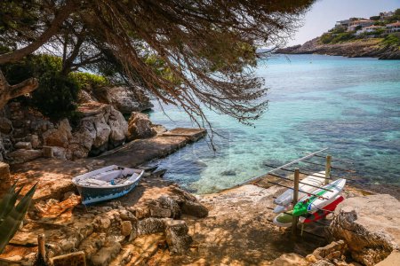 A serene view of Font de Sa Cala beach in Mallorca, featuring a small boat and paddleboards resting on a rocky shoreline. The clear turquoise waters are surrounded by natural rock formations and lush greenery