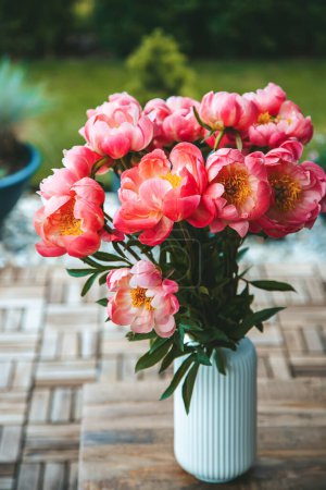 A beautiful bouquet of vibrant pink peonies with lush petals. Flowers are housed in a white vase with vertical ridging, placed on a wooden table
