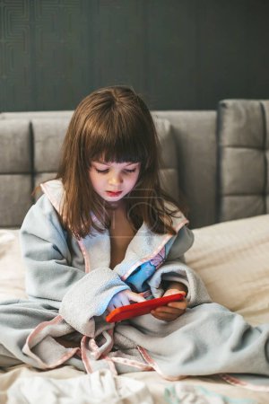 A young girl wearing a bathrobe is deeply engrossed in her smartphone. Concept: kids and smartphones