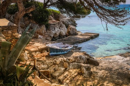 Tranquil scene of Font de Sa Cala beach in Mallorca, showcasing a lone boat nestled on the rocky coast amidst turquoise waters and verdant foliage