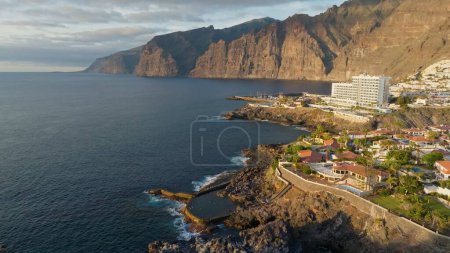 Téléchargez les photos : Aerial view of Los Gigantes restort on Tenerife Canary island. Flying over magnificent hotels, villas and natural pool on the ocean - rocks in sunset light in the background. Canary islands, Spain - en image libre de droit