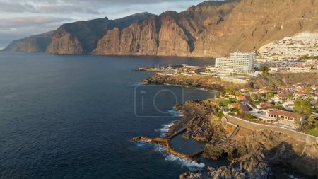 Téléchargez les photos : Aerial view of Los Gigantes restort on Tenerife Canary island. Flying over magnificent hotels, villas and natural pool on the ocean - rocks in sunset light in the background. Canary islands, Spain - en image libre de droit