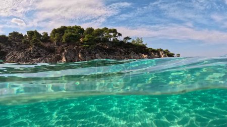 Photo for Crystal clear sea water in Sitonia, Halkidiki, Greece. Shot from border of air and water with dome. Half underwater slow-motion view of sandy beach bottom and rock with trees 2 - Royalty Free Image