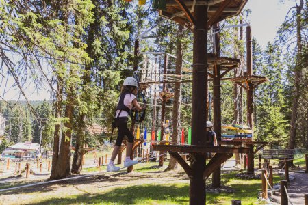 Téléchargez les photos : Jeune fille intrépide dans un parc d'aventure au sommet d'un arbre au cœur d'une forêt luxuriante pendant un camp d'été. - en image libre de droit
