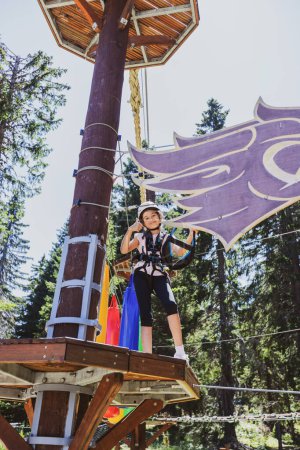 Téléchargez les photos : Joyeux enfant fille au parc d'aventure en forêt sur le camp d'été, portant des engins de protection. - en image libre de droit