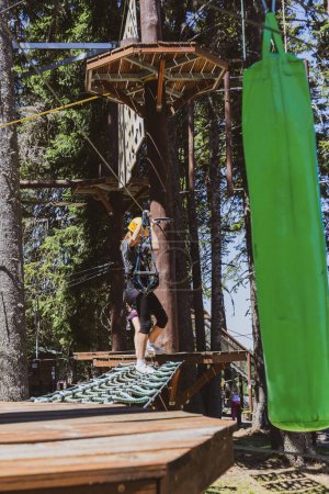 Foto de Chica cruza un curso de cuerda desafiante en un bosque en el parque de atracciones, usando equipo de protección. - Imagen libre de derechos