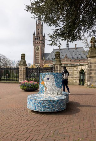 Téléchargez les photos : La Haye, Pays-Bas - 17 avril 2023 : Banc avec une colombe blanche tenant une branche d'olivier symbole de paix devant le Palais de la Paix de La Haye, qui abrite la Cour internationale de justice - en image libre de droit