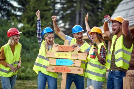 Photo for A group of cheerful both female and male builders posing for a photo at a cottage construction site on a beautiful day. Construction, building, workers - Royalty Free Image