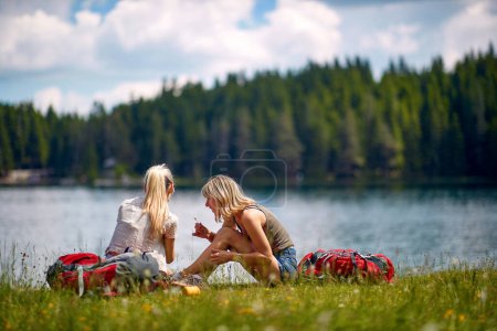 Téléchargez les photos : Deux copines caucasiennes assises au bord du lac, faisant une pause de randonnée, parlant, souriant - en image libre de droit