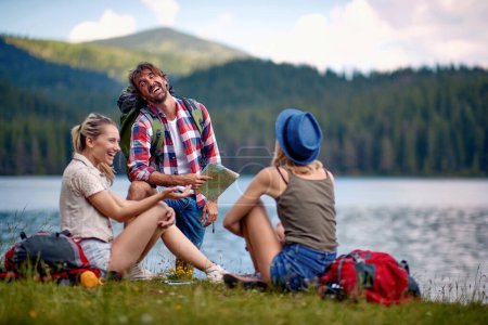 Foto de Grupo de tres, hombre y dos mujeres, tomando un descanso en el lago, hablando, riendo. estilo de vida saludable - Imagen libre de derechos