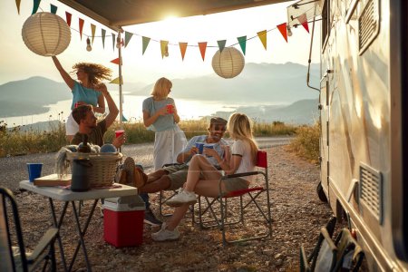 Photo for Group of young people socializing, talking, smiling, dancing in front of a camper with decorated awning - Royalty Free Image