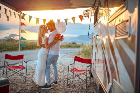 Téléchargez les photos : Jeune couple adulte étreignant, se tenant dans les bras, se regardant en amour, devant un camping-car en plein air au coucher du soleil et belle vue - en image libre de droit