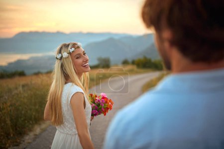 Photo for Portrait of beautiful blonde turned around in motion with red bouquet in her hands and  of flowers on her head, smiling, laughing, looking at her fiance - Royalty Free Image