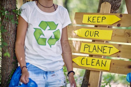 Foto de Mujer con bolsa de plástico para recoger basura, de pie delante de un tablero de madera diciendo: Mantenga nuestro planeta limpio. Ambientalismo, concepto de estilo de vida. - Imagen libre de derechos