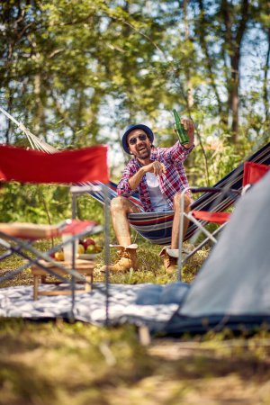 Photo for Handsome young hipster male cheering with beer and sitting in hammock. Holiday, leisure, fun, lifestyle concept. - Royalty Free Image