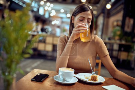 Téléchargez les photos : Jolie jeune femme assise seule dans un café dégustant son jus et son morceau de gâteau. Vie étudiante, jeune adulte, mode de vie. - en image libre de droit