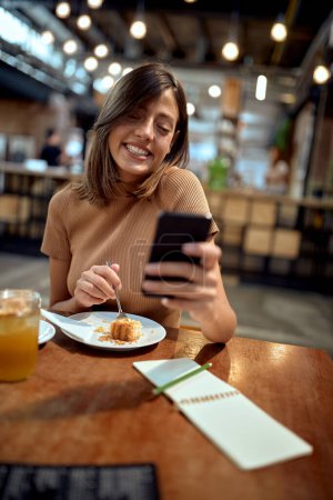 Photo for Young woman at coffee shop, looking at phone and smiling. Cheerful happy girl enjoying her time. Student life, young adult, lifestyle. - Royalty Free Image