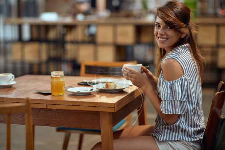 Foto de Mujer joven con hermosa sonrisa sosteniendo la taza de café y sintiéndose alegre. Mujer con lápiz y papel y auriculares en la cafetería. - Imagen libre de derechos