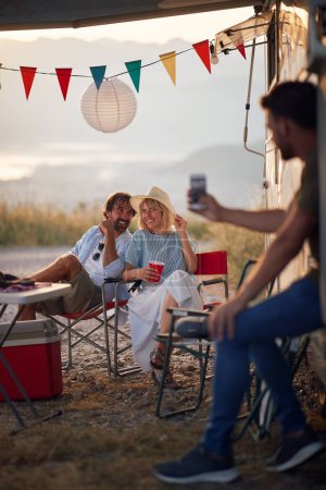 Téléchargez les photos : Un groupe d'amis assis au camping dans la nature et aime bavarder et boire par une belle journée. Amitié, vacances, camping, nature - en image libre de droit
