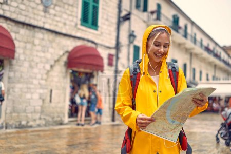 Foto de Mujer joven en impermeable en la calle con mapa, sintiéndose alegre. Lluvia de verano. Viajes, concepto de estilo. - Imagen libre de derechos