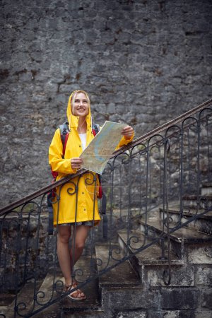 Foto de Mujer con impermeable sosteniendo mapa, sintiéndose alegre. Lluvia de verano. Viajes, ciudad, concepto de estilo de vida. - Imagen libre de derechos