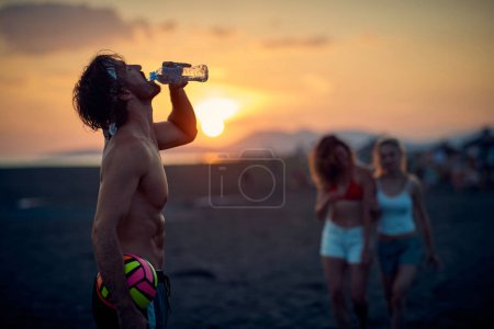 Téléchargez les photos : Homme avec volley-ball à la plage boire de l'eau, au coucher du soleil. Je me prépare à jouer au volley avec des amis. Sport, plaisir, convivialité, concept de style de vie. - en image libre de droit