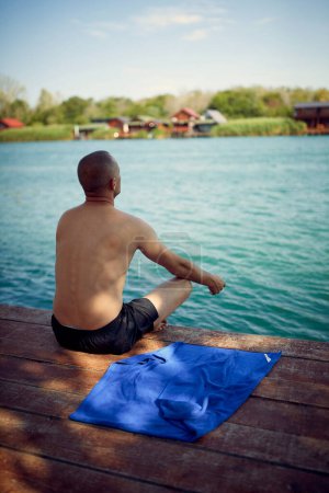 Photo for Man doing morning meditation by water. Young man sitting on wooden jetty by sea doing spiritual practice. Relaxation, discipline, lifestyle concept. - Royalty Free Image