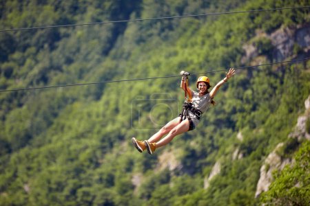 Photo for Happy young cheerful female tourist wearing casual clothes riding on zipline in forest.  Zipline trip selective focus against blurred background. Holiday, adventure, extreme sport concept. - Royalty Free Image
