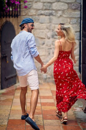 Photo for Beautiful young couple holding hands and running together. Beautiful woman in red summer dress, stylish handsome man. Date, love, holiday concept. - Royalty Free Image