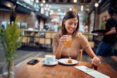 Photo for Cheerful young woman in coffee shop, enjoying her breakfast and coffee while taking notes. Student life, young adult, lifestyle. - Royalty Free Image