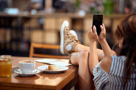 Téléchargez les photos : Jeune femme avec téléphone portable assis à table avec les jambes levées, écoutant des écouteurs, profitant de son temps dans un café. - en image libre de droit