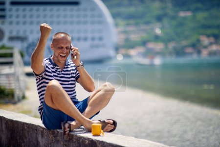 Foto de Un joven está hablando en el teléfono inteligente mientras descansa en el muelle en un hermoso día soleado durante unas vacaciones en la playa. Vacaciones, playa, turistas - Imagen libre de derechos