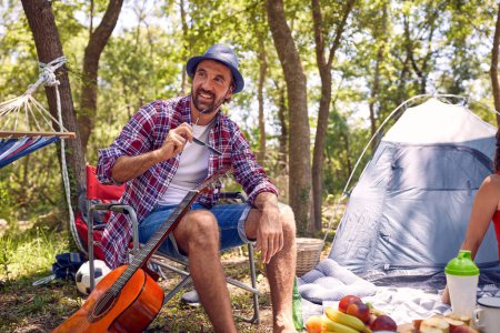 Foto de Joven hipster hombre sonriendo, sentado en la silla y disfrutando de acampar en la naturaleza. Picnic en el bosque. Vacaciones, ocio, estilo de vida. - Imagen libre de derechos