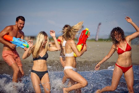 Téléchargez les photos : Groupe d'amis jouant avec des pistolets à eau dans l'eau à la plage. Jeunes adultes en vacances d'été ensemble. Vacances, plaisir, concept de style de vie. - en image libre de droit