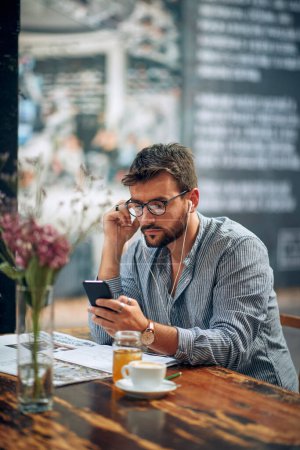 Téléchargez les photos : Beau jeune homme avec des lunettes en tenue décontractée en utilisant un smartphone avec écouteurs, profiter de la pause café seul. Interieur de café moderne. - en image libre de droit