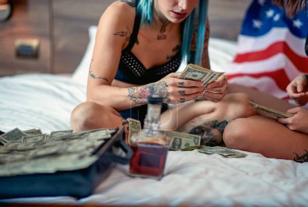 Téléchargez les photos : Un jeune couple lesbien tatoué en lingerie minutieuse comptant a gagné de l'argent dans une atmosphère détendue dans la chambre. Love, lgbt, USA, luxe, relation - en image libre de droit