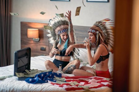 Photo for A cheerful sexy tattooed lesbian couple in the lingerie and with indian headdress is sitting on the bed while is drinking whisky and celebrating by throwing a bunch of money in the air in a festive atmosphere. Home, relationship, love - Royalty Free Image